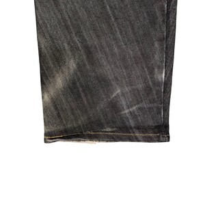 Op de afbeelding: Close-up van de zoom van een donkergrijze denim jeans. De stof heeft een textuur met lichtere plekken, wat slijtage suggereert. De zoom is afgewerkt met geelgouden stiksels.