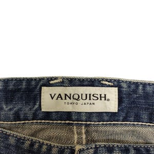 May include: Close-up of blue denim jeans with a white label that reads "VANQUISH TOKYO JAPAN". The jeans have a button closure and visible stitching.