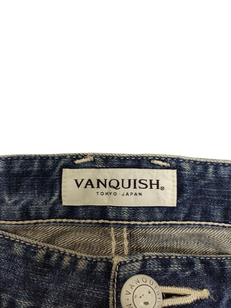 May include: Close-up of blue denim jeans with a white label that reads "VANQUISH TOKYO JAPAN". The jeans have a button closure and visible stitching.