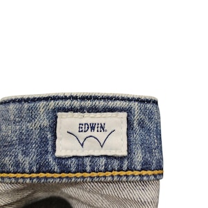 May include: Close-up of a denim jeans waistband. A white tag with the brand name "EDWIN" and a stylized wave logo is stitched onto the blue denim. The jeans have yellow stitching.
