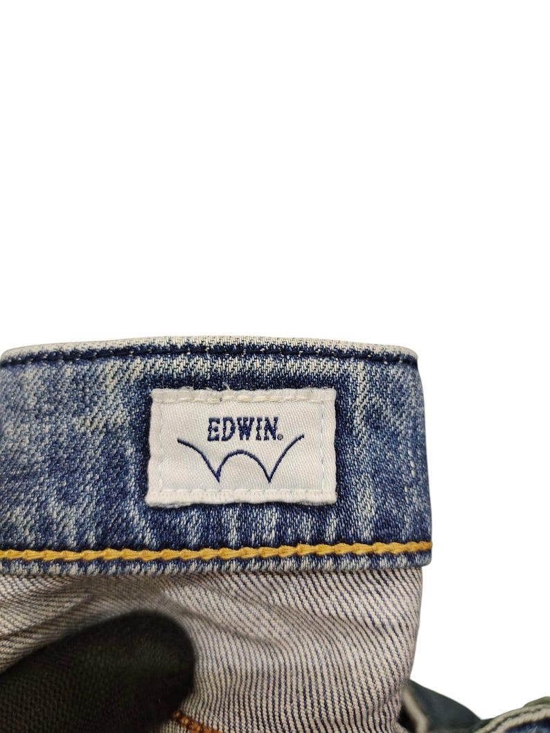 May include: Close-up of a denim jeans waistband. A white tag with the brand name "EDWIN" and a stylized wave logo is stitched onto the blue denim. The jeans have yellow stitching.