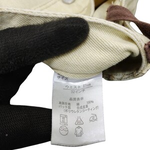 Puede incluir: Primer plano de unos pantalones vaqueros color crema. La cintura mide 81 cm (32 pulgadas). Una etiqueta con texto japon&eacute;s detalla la composici&oacute;n del material y las instrucciones de cuidado. Los pantalones son 100% algod&oacute;n.