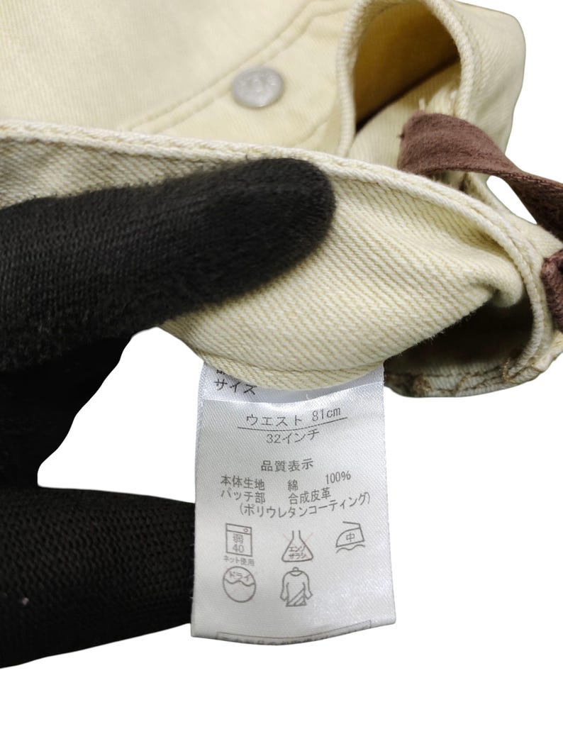 Puede incluir: Primer plano de unos pantalones vaqueros color crema. La cintura mide 81 cm (32 pulgadas). Una etiqueta con texto japon&eacute;s detalla la composici&oacute;n del material y las instrucciones de cuidado. Los pantalones son 100% algod&oacute;n.