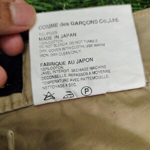 Puede incluir: Una etiqueta blanca con texto negro unida a una prenda beige. La etiqueta dice "COMME des GAR&Ccedil;ONS Co., Ltd. TC-P025 MADE IN JAPAN 100% COTTON DO NOT BLEACH. DO NOT TUMBLE DRY. COVER WITH CLOTH, USE WARM IRON. DRY CLEAN ONLY. FABRIQUE AU JAPON 100% COTON JAVEL INTERDIT. SECHAGE MACHINE DECONSEILLE. REPASSER A MOYENNE TEMPERATURE AVEC PATTEMOUILLE. NETTOYAGE A SEC."