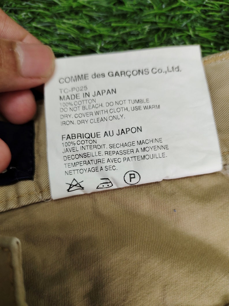 Puede incluir: Una etiqueta blanca con texto negro unida a una prenda beige. La etiqueta dice "COMME des GAR&Ccedil;ONS Co., Ltd. TC-P025 MADE IN JAPAN 100% COTTON DO NOT BLEACH. DO NOT TUMBLE DRY. COVER WITH CLOTH, USE WARM IRON. DRY CLEAN ONLY. FABRIQUE AU JAPON 100% COTON JAVEL INTERDIT. SECHAGE MACHINE DECONSEILLE. REPASSER A MOYENNE TEMPERATURE AVEC PATTEMOUILLE. NETTOYAGE A SEC."