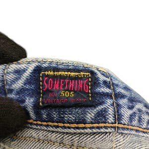 Puede incluir: Primer plano de una etiqueta de jeans de mezclilla con el texto "SOMETHING" en rojo y amarillo, "LOT 505 VINTAGE JEANS" en blanco. La etiqueta est&aacute; cosida sobre tela de mezclilla azul.