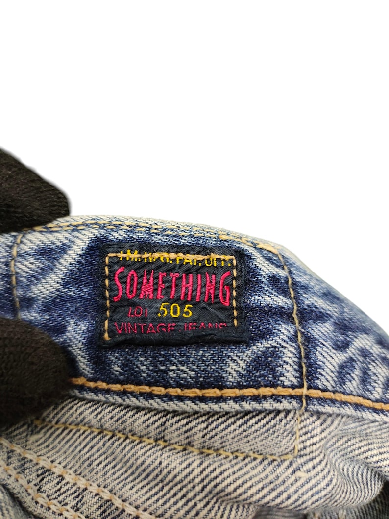 Puede incluir: Primer plano de una etiqueta de jeans de mezclilla con el texto "SOMETHING" en rojo y amarillo, "LOT 505 VINTAGE JEANS" en blanco. La etiqueta est&aacute; cosida sobre tela de mezclilla azul.