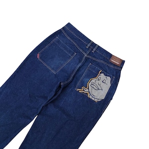 May include: Dark blue denim jeans featuring a cartoon face design on the back pocket. The jeans have a classic five-pocket design and a brown leather patch. The jeans are made of a durable denim material.