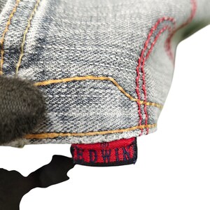 May include: Close-up of denim fabric with red stitching and a red tag that reads "EDWIN" in white lettering. The denim is a light blue wash with yellow stitching.