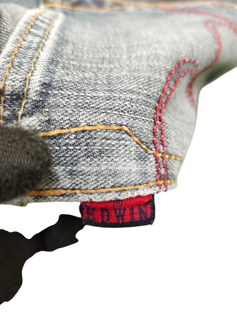 May include: Close-up of denim fabric with red stitching and a red tag that reads "EDWIN" in white lettering. The denim is a light blue wash with yellow stitching.