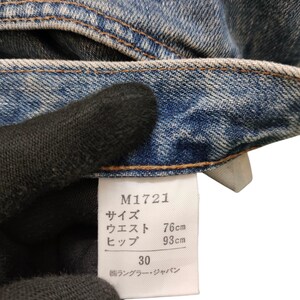 May include: Close-up of blue denim jeans with a white tag. The tag displays the product code M1721, waist size 30, and measurements of 76cm waist and 93cm hips. The jeans are made of a durable denim fabric.