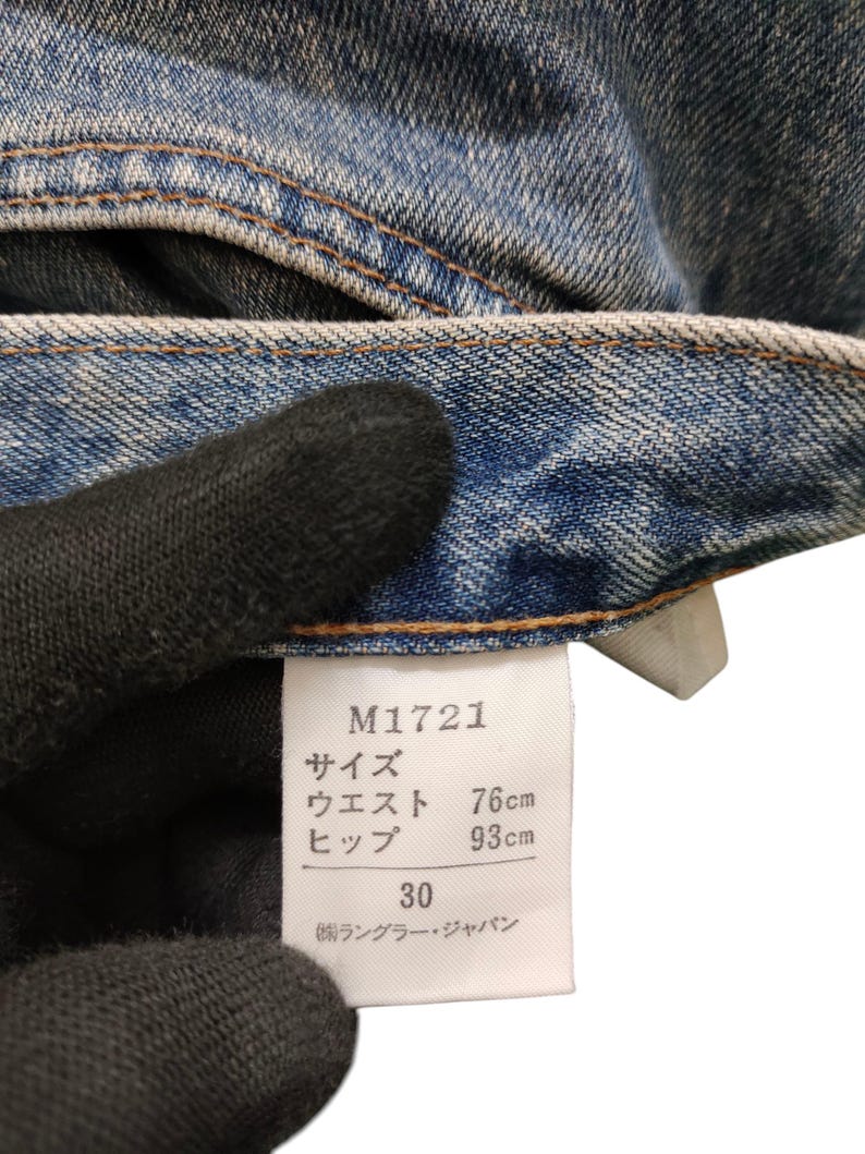 May include: Close-up of blue denim jeans with a white tag. The tag displays the product code M1721, waist size 30, and measurements of 76cm waist and 93cm hips. The jeans are made of a durable denim fabric.