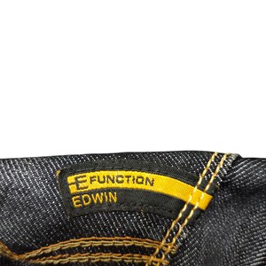 Op de afbeelding: Close-up van donkere denimstof met een geel en zwart label. Het label leest "E FUNCTION EDWIN" in gele en witte tekst. De stiksels zijn in een contrasterende goudkleur.