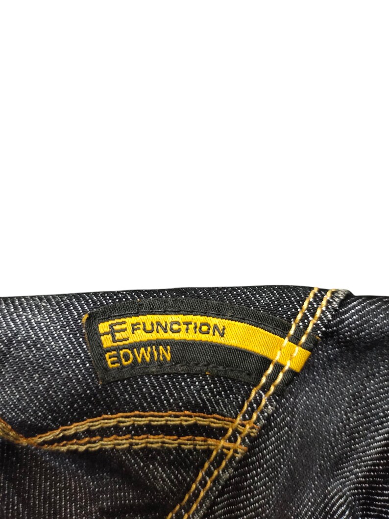 Op de afbeelding: Close-up van donkere denimstof met een geel en zwart label. Het label leest "E FUNCTION EDWIN" in gele en witte tekst. De stiksels zijn in een contrasterende goudkleur.