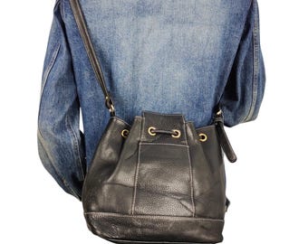 May include: Black leather bucket bag with a drawstring closure and a shoulder strap. The bag has gold-toned metal eyelets and a stitched design. The bag is worn over a denim jacket.