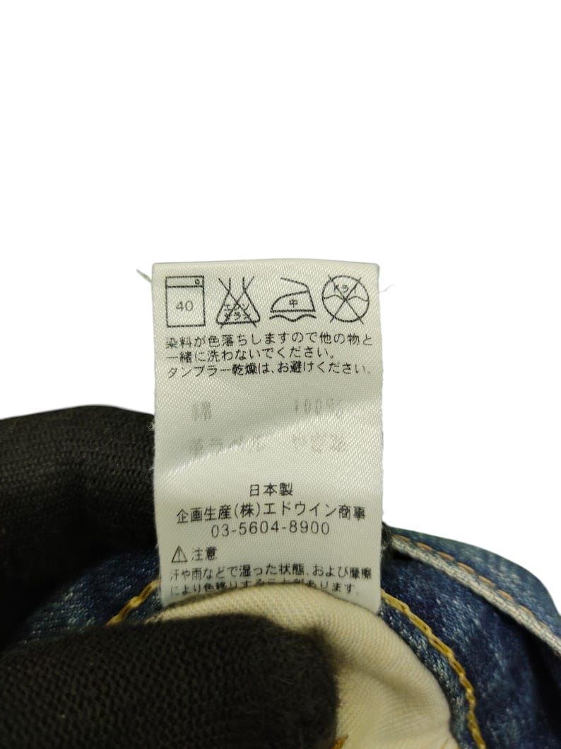 Puede incluir: Primer plano de una etiqueta blanca con texto japon&eacute;s y s&iacute;mbolos de cuidado. La etiqueta incluye instrucciones de lavado, una composici&oacute;n de tejido del 100% e informaci&oacute;n del fabricante. La etiqueta est&aacute; adherida a unos vaqueros azules.