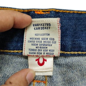 K&ouml;nnte beinhalten: Ein wei&szlig;es Pflegeetikett mit schwarzem Text, das an einer blauen Jeans befestigt ist. Das Etikett lautet "RN#112790 CA#30427 100% BAUMWOLLE MASCHINENW&Auml;SCHE KALT SCHONWASCHGANG AUF LINKS WASCHEN SEPARAT KEIN BLEICHMITTEL TROCKNEN IM TROCKNER BEI SCHWACHER HITZE WARM B&Uuml;GELN, WENN ERFORDERLICH" und zeigt ein rotes True Religion Hufeisen-Logo.