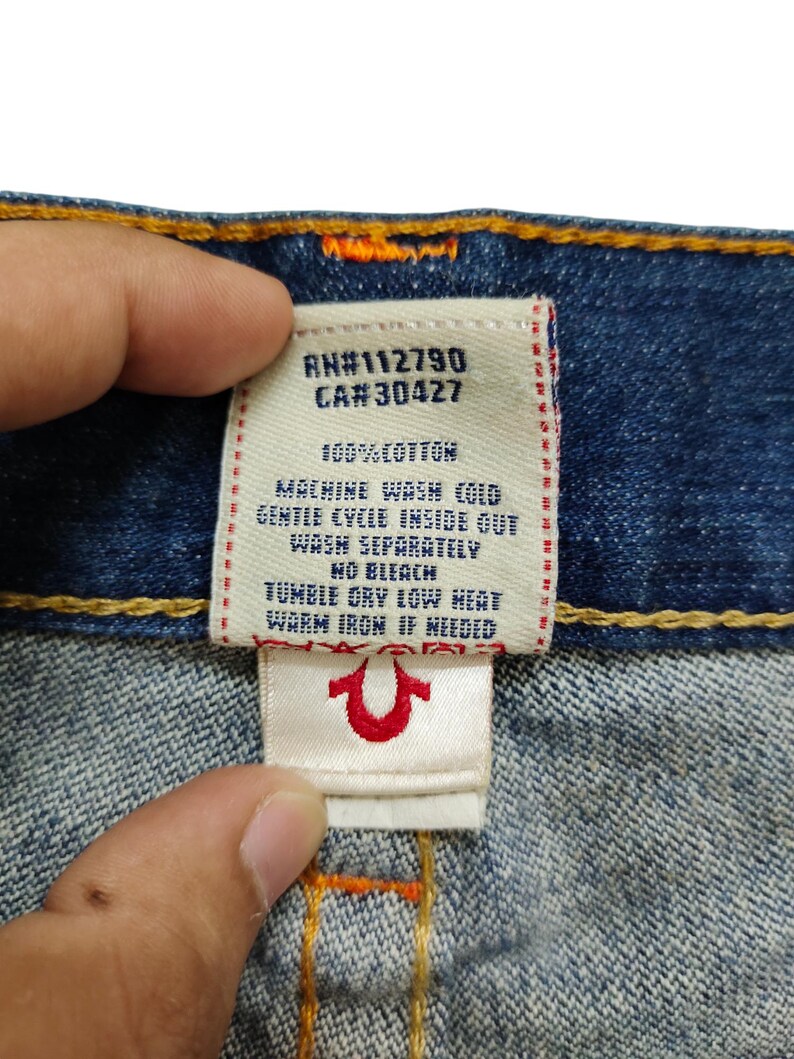 K&ouml;nnte beinhalten: Ein wei&szlig;es Pflegeetikett mit schwarzem Text, das an einer blauen Jeans befestigt ist. Das Etikett lautet "RN#112790 CA#30427 100% BAUMWOLLE MASCHINENW&Auml;SCHE KALT SCHONWASCHGANG AUF LINKS WASCHEN SEPARAT KEIN BLEICHMITTEL TROCKNEN IM TROCKNER BEI SCHWACHER HITZE WARM B&Uuml;GELN, WENN ERFORDERLICH" und zeigt ein rotes True Religion Hufeisen-Logo.