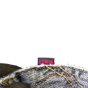 May include: Close-up of a section of blue denim jeans with visible stitching and a small rectangular tag. The tag is navy blue with the brand name "EDWIN" in red lettering.