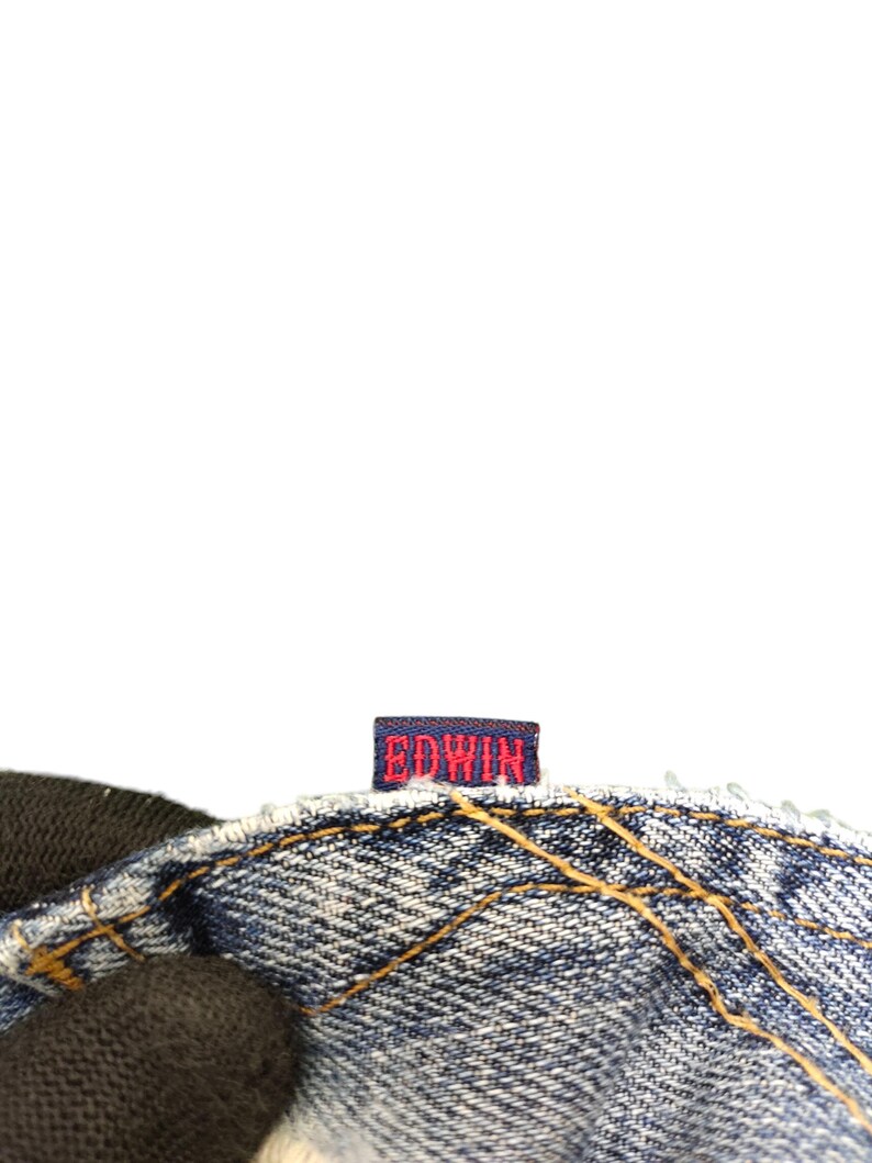 May include: Close-up of a section of blue denim jeans with visible stitching and a small rectangular tag. The tag is navy blue with the brand name "EDWIN" in red lettering.