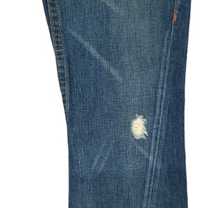 K&ouml;nnte beinhalten: Nahaufnahme von blauem Jeansstoff mit einem kleinen wei&szlig;en Loch. Der Stoff hat einen abgenutzten und verblassten Look mit sichtbaren N&auml;hten an den R&auml;ndern.