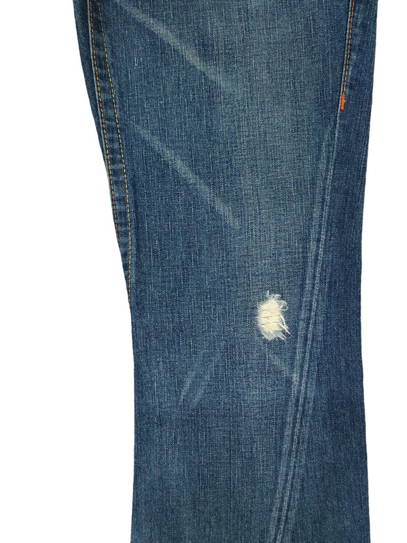 K&ouml;nnte beinhalten: Nahaufnahme von blauem Jeansstoff mit einem kleinen wei&szlig;en Loch. Der Stoff hat einen abgenutzten und verblassten Look mit sichtbaren N&auml;hten an den R&auml;ndern.