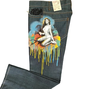 May include: Blue denim jeans with a colorful graphic print of a woman in a white swimsuit. The print features a spray paint effect with dripping paint.