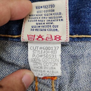 May include: A blue denim jean label with white text and red stitching. The label includes the following information: RN#112790, 100% Cotton, Machine Wash Cold, Tumble Dry Medium, Color May Transfer When New, Wash Inside Out To Retain Color, Cut#600137, Style#04-803, P.O#50591, 100% Cotton, 100% Coton, Made in USA, Fabrique au EUA, CA#30422.