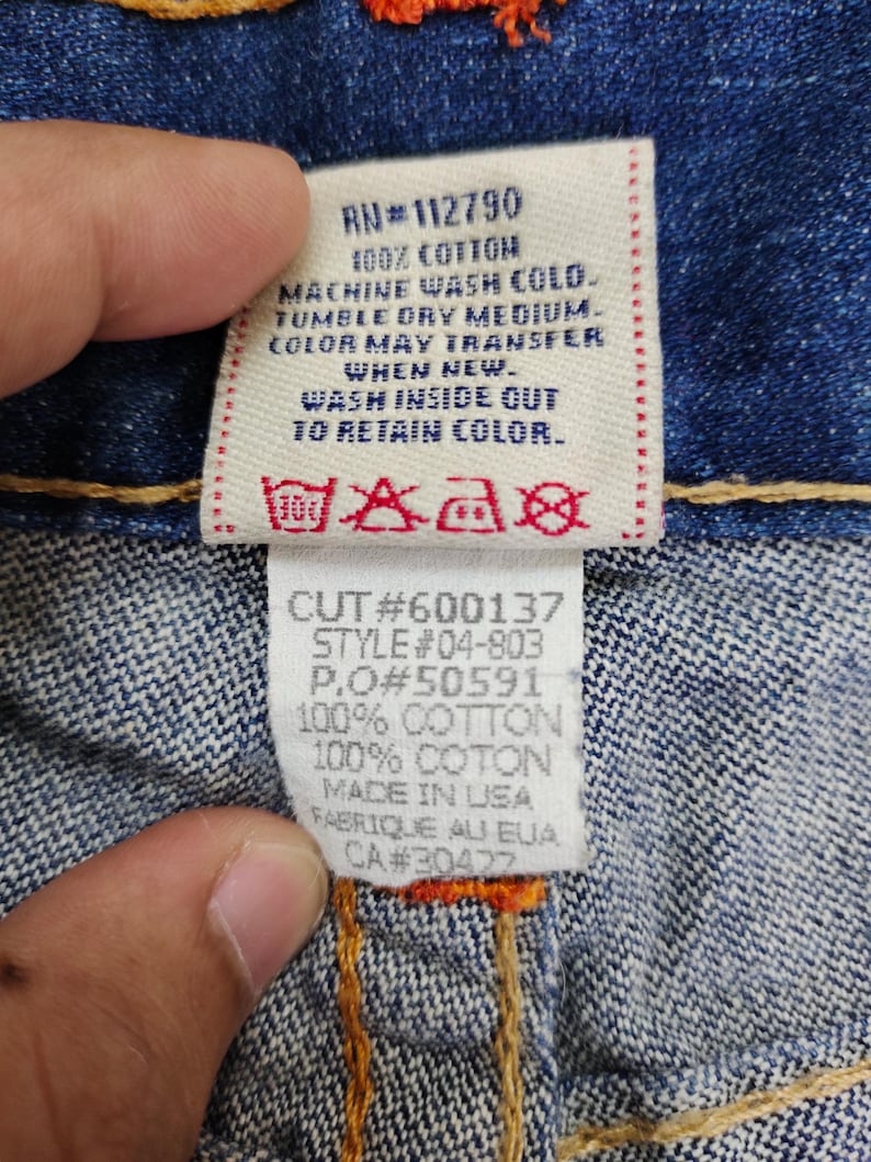 May include: A blue denim jean label with white text and red stitching. The label includes the following information: RN#112790, 100% Cotton, Machine Wash Cold, Tumble Dry Medium, Color May Transfer When New, Wash Inside Out To Retain Color, Cut#600137, Style#04-803, P.O#50591, 100% Cotton, 100% Coton, Made in USA, Fabrique au EUA, CA#30422.