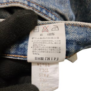 May include: Close-up of a denim garment with a white tag. The tag features Japanese text, care symbols, and the text "NIP". Below the text are several lines of numbers and letters. The denim is a light blue color.