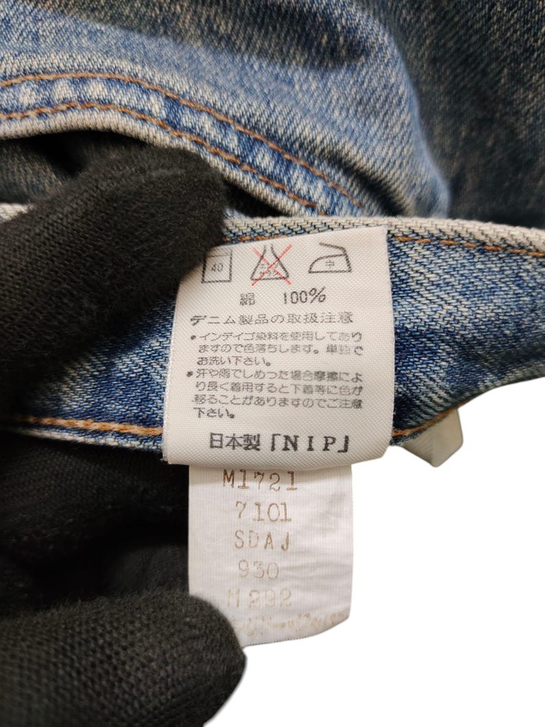 May include: Close-up of a denim garment with a white tag. The tag features Japanese text, care symbols, and the text "NIP". Below the text are several lines of numbers and letters. The denim is a light blue color.
