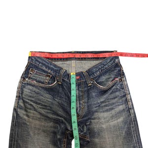 May include: Blue denim jeans with a faded wash, featuring a red measuring tape across the waist and a green measuring tape down the inseam. The jeans have classic five-pocket styling and a button fly.