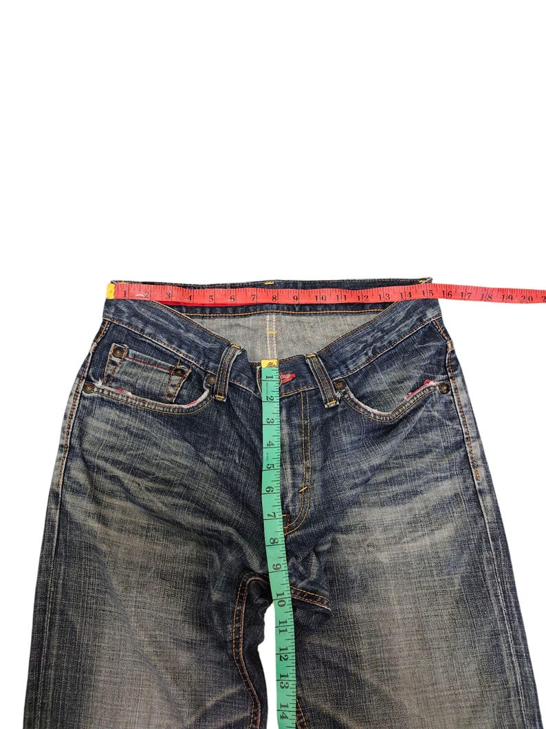 May include: Blue denim jeans with a faded wash, featuring a red measuring tape across the waist and a green measuring tape down the inseam. The jeans have classic five-pocket styling and a button fly.