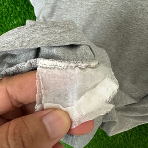 Może przedstawiać: Zbliżenie na szary t-shirt z przymocowaną białą metką z tkaniny. Metka ma obszyty brzeg.