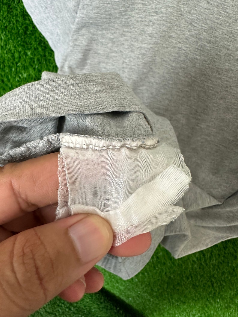 Może przedstawiać: Zbliżenie na szary t-shirt z przymocowaną białą metką z tkaniny. Metka ma obszyty brzeg.