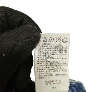 May include: Close-up of a white tag with Japanese text and care symbols attached to a pair of blue denim jeans. The tag includes the number 34. The jeans are partially visible, with a dark blue wash.