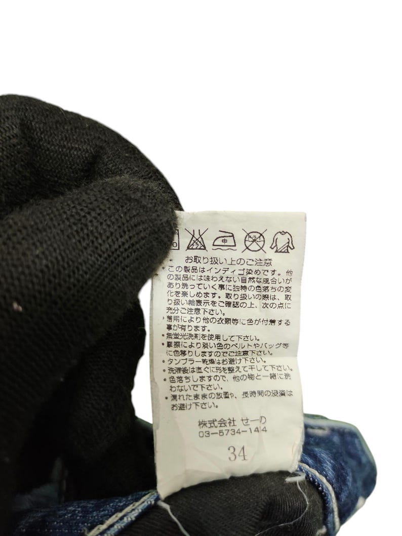 May include: Close-up of a white tag with Japanese text and care symbols attached to a pair of blue denim jeans. The tag includes the number 34. The jeans are partially visible, with a dark blue wash.