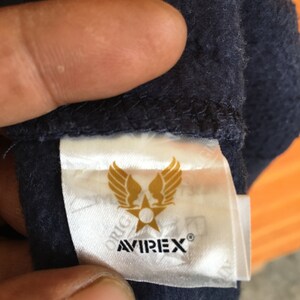 May include: A white tag with a gold star and wings logo. The text "AVIREX&reg;" is printed in black below the logo. The tag is attached to a dark blue fabric.