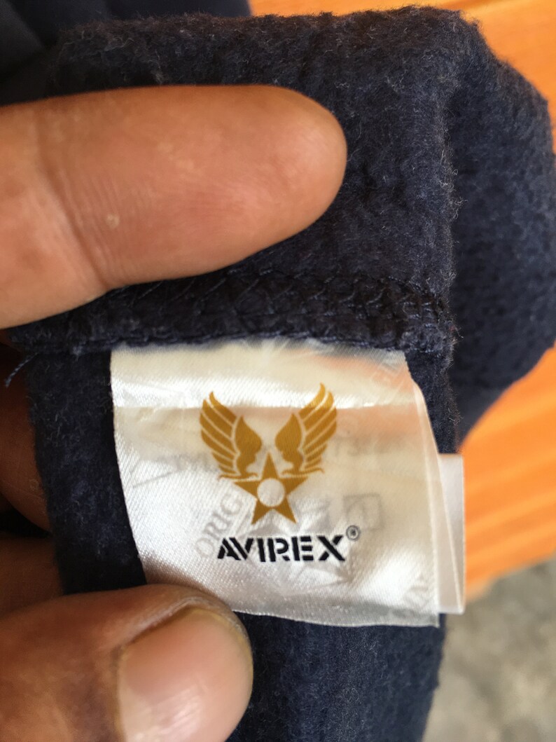 May include: A white tag with a gold star and wings logo. The text "AVIREX&reg;" is printed in black below the logo. The tag is attached to a dark blue fabric.
