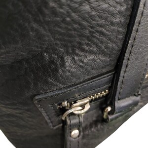 May include: Close-up of a black leather bag with a silver zip and a silver buckle. The bag has a textured surface.