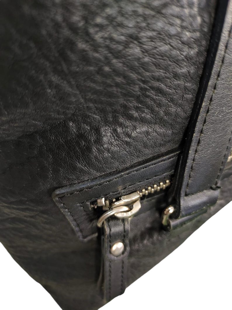 May include: Close-up of a black leather bag with a silver zip and a silver buckle. The bag has a textured surface.