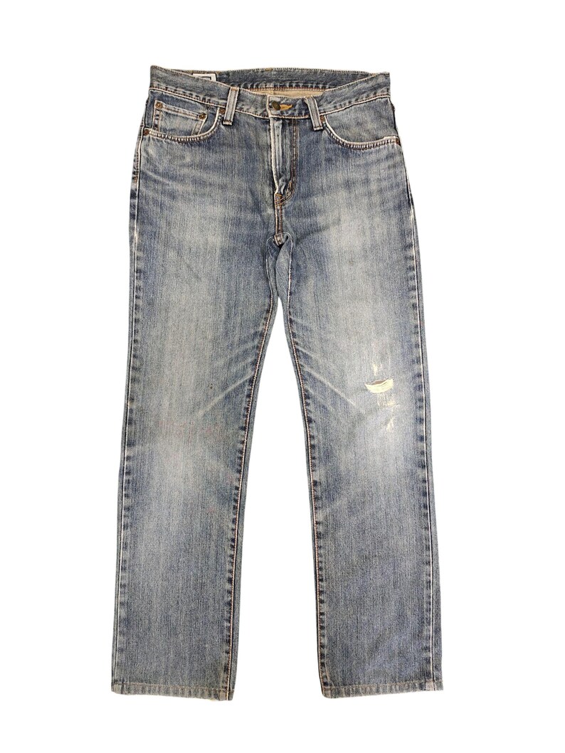 May include: Light blue denim jeans with a classic five-pocket design. The jeans have a faded wash and a distressed area on the right leg. The jeans are straight-legged and have a button and zipper closure.