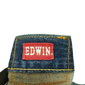 Puede incluir: Primer plano de una etiqueta de jeans de mezclilla con el nombre de la marca "EDWIN" en letras blancas sobre un parche rectangular rojo. Los jeans tienen una tela de mezclilla azul con costuras amarillas.
