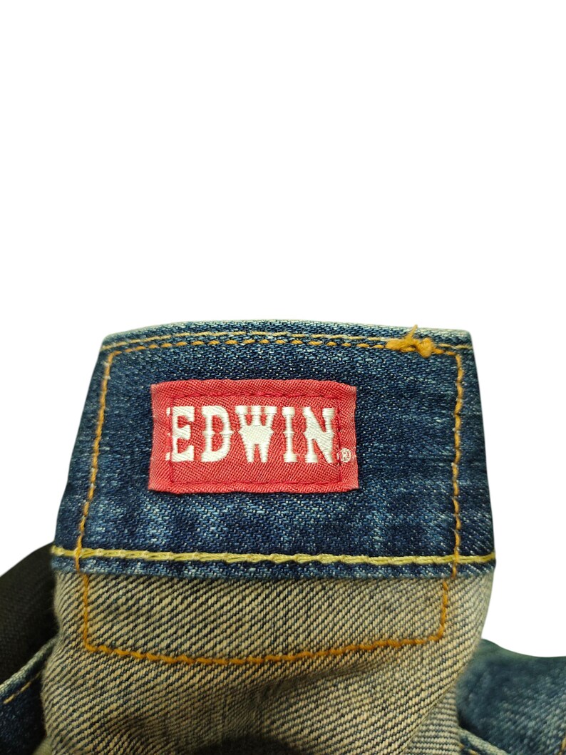Puede incluir: Primer plano de una etiqueta de jeans de mezclilla con el nombre de la marca "EDWIN" en letras blancas sobre un parche rectangular rojo. Los jeans tienen una tela de mezclilla azul con costuras amarillas.