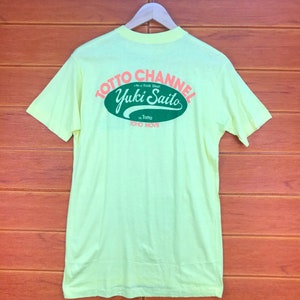 May include: Yellow t-shirt with a green and orange logo that says "TOTO CHANNEL - A film of Kazuki Omori - Yuki Saito in TOTO - TOHO MOVIE"