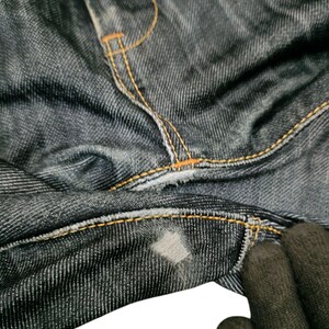 Op de afbeelding: Close-up van een donkerblauwe denim jeans met oranje stiksels. De jeans vertoont slijtage, met een zichtbare patch en gerafelde randen. De stof heeft een getextureerd uiterlijk.