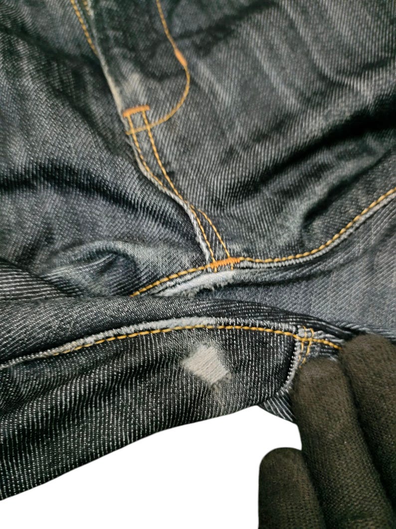 Op de afbeelding: Close-up van een donkerblauwe denim jeans met oranje stiksels. De jeans vertoont slijtage, met een zichtbare patch en gerafelde randen. De stof heeft een getextureerd uiterlijk.