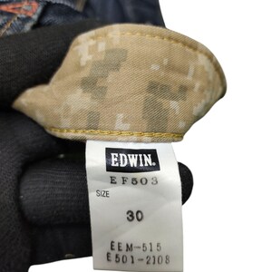 Op de afbeelding: Close-up van een denim kledinglabel met de merknaam "EDWIN". Het label toont "EF503" en "MAAT 30". De binnenkant van het kledingstuk heeft een camouflagepatroon. Het label bevat ook de nummers "EEM-515" en "E501-2108".