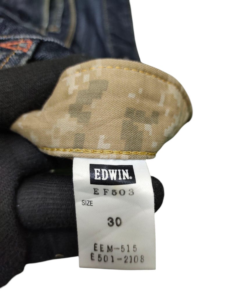 Op de afbeelding: Close-up van een denim kledinglabel met de merknaam "EDWIN". Het label toont "EF503" en "MAAT 30". De binnenkant van het kledingstuk heeft een camouflagepatroon. Het label bevat ook de nummers "EEM-515" en "E501-2108".