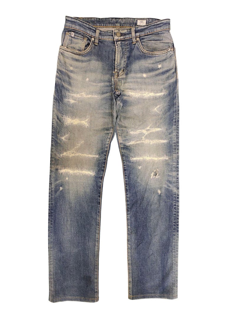 May include: Faded blue denim jeans with distressed detailing. The jeans feature a classic five-pocket design, a button and zipper closure, and visible wear and tear throughout the fabric. The jeans have a straight leg cut.